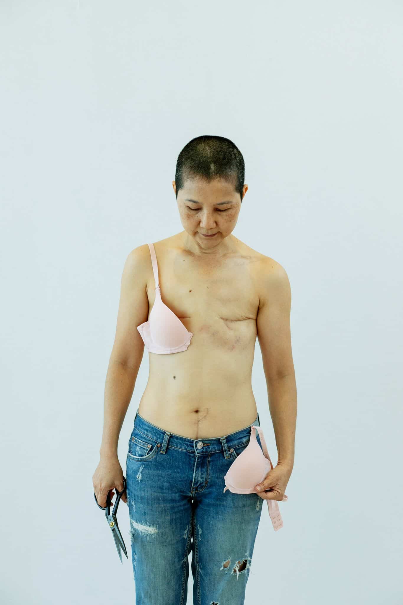 Forside Serious mature ethnic female in jeans with cut bra and scissors in hand after breast removal surgery from cancer standing in light room on white background